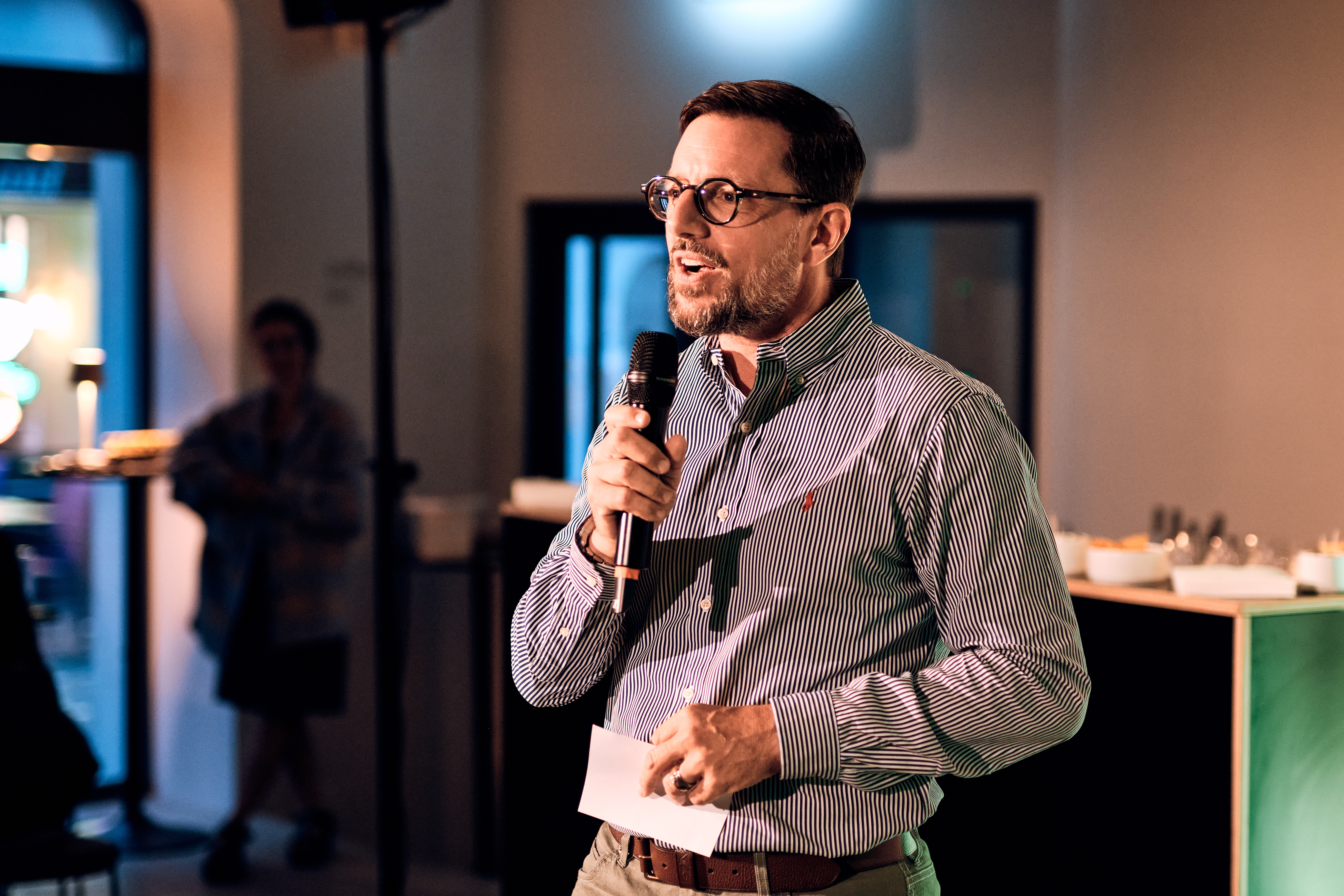 Ein Mann mit Brille und gestreiftem Hemd spricht bei einer Indoor-Veranstaltung in ein Mikrofon und hält Papier in der Hand.